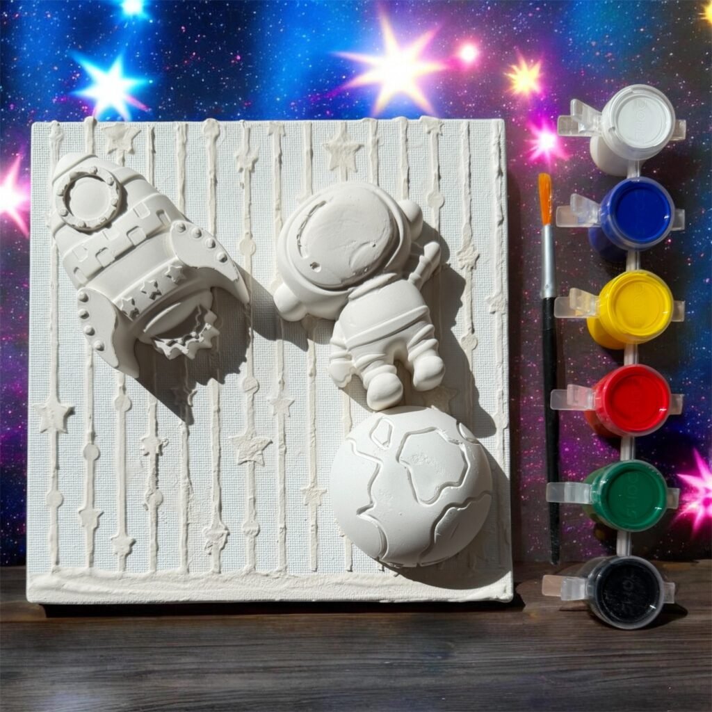 A 6x6 inch DIY space-themed art kit. On a white, textured canvas, there are three 3D plaster sculptures: a rocket ship, an astronaut, and a planet. To the right is a black paintbrush and a strip of six paint pots (white, blue, yellow, red, green, black). The background features a vibrant, colorful galaxy and star pattern.