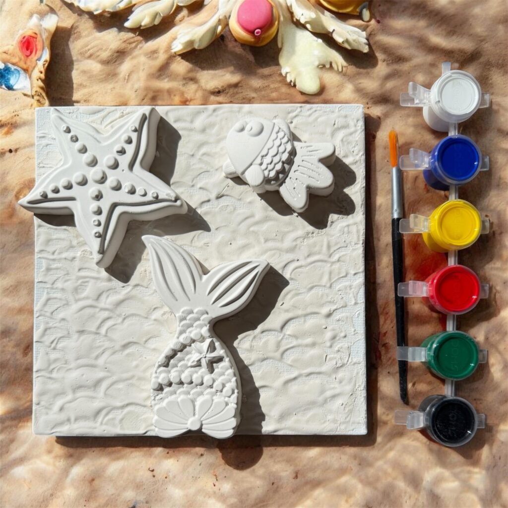 A top-down view of a DIY mixed media art kit. It features a square white canvas with a wavy texture, adorned with three 3D plaster shapes: a starfish, a mermaid tail, and a fish. To the right, there is a paintbrush and a strip of six acrylic paint pots in white, blue, yellow, red, green, and black. The kit is set against a sandy, sun-drenched background with beach-themed decor at the top.