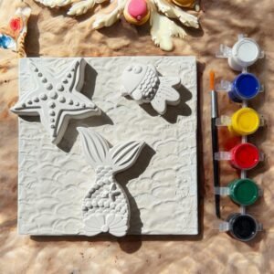 A top-down view of a DIY mixed media art kit. It features a square white canvas with a wavy texture, adorned with three 3D plaster shapes: a starfish, a mermaid tail, and a fish. To the right, there is a paintbrush and a strip of six acrylic paint pots in white, blue, yellow, red, green, and black. The kit is set against a sandy, sun-drenched background with beach-themed decor at the top.