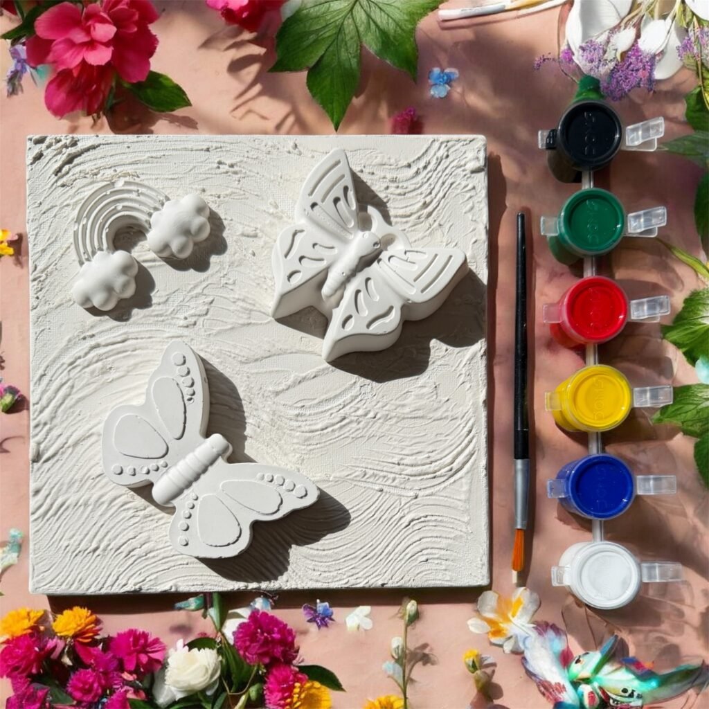 A 6x6 inch mixed media art kit featuring a white textured canvas with three 3D plaster shapes: two butterflies and a rainbow with clouds. To the right is a professional paintbrush and a strip of six acrylic paint pots in black, green, red, yellow, blue, and white. The scene is framed by vibrant pink and yellow flowers and green leaves on a soft pink background.