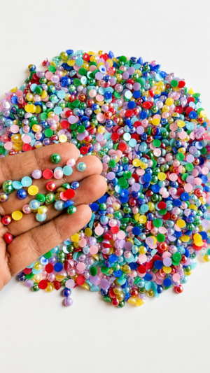 A 100g pile of vibrant, multi-colored flatback half-pearls spread on a white surface, with a hand holding a small handful of the iridescent, glossy craft beads to showcase their size and shiny finish.