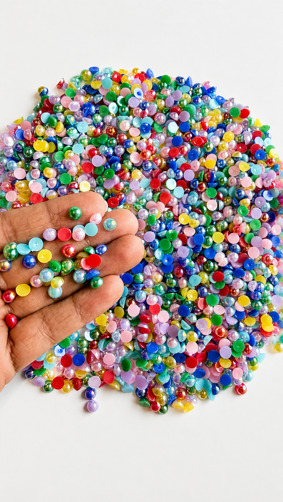 A 100g pile of vibrant, multi-colored flatback half-pearls spread on a white surface, with a hand holding a small handful of the iridescent, glossy craft beads to showcase their size and shiny finish.