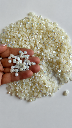 A 100g mound of glossy ivory, off-white flatback half-pearls spread on a clean white surface. A hand lifts a small handful of the beads close to the camera, showcasing their smooth, flat bottoms and the subtle, rainbow-colored iridescent AB finish that reflects off their round tops.