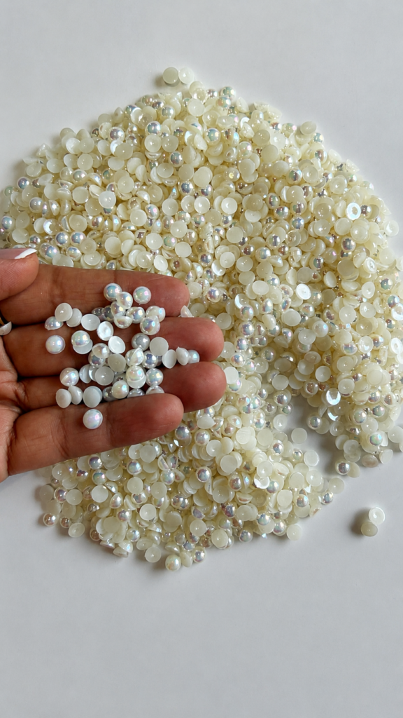 A 100g mound of glossy ivory, off-white flatback half-pearls spread on a clean white surface. A hand lifts a small handful of the beads close to the camera, showcasing their smooth, flat bottoms and the subtle, rainbow-colored iridescent AB finish that reflects off their round tops.