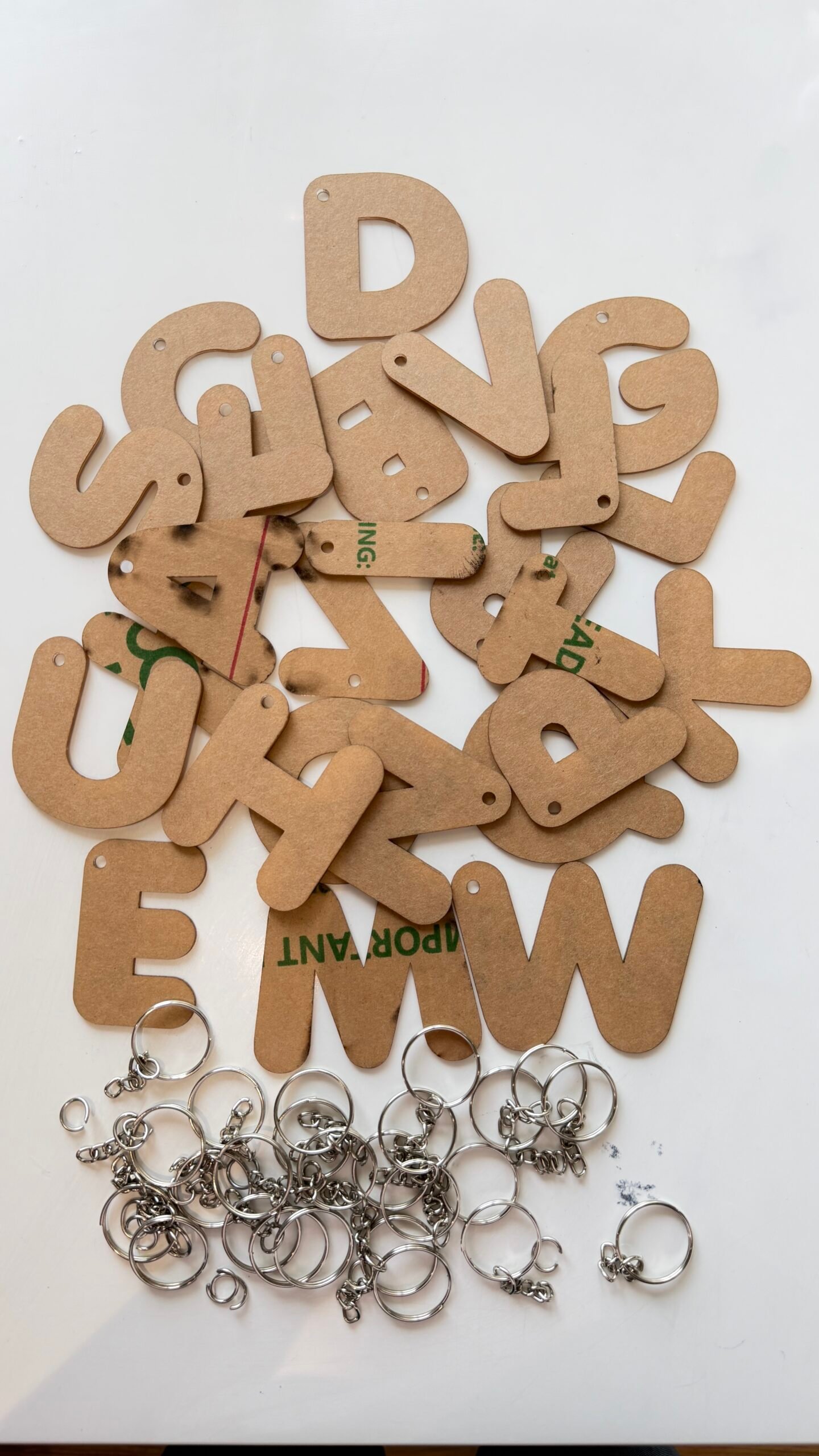 A 52-piece DIY craft kit featuring 26 alphabet letter blanks from A to Z covered in brown protective paper, arranged on a white background above a pile of 26 silver metal keychain rings and chains.