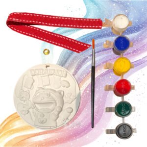 An all-in-one DIY craft hanging kit arranged against a whimsical colorful background. The kit features a white, 4-inch round embossed keepsake plate with a 3D Doraemon character design, a fine-tip paintbrush, a strip of six vibrant acrylic paint pots, a red ribbon with white contrast stitching, and a single golden metal eyelet.
