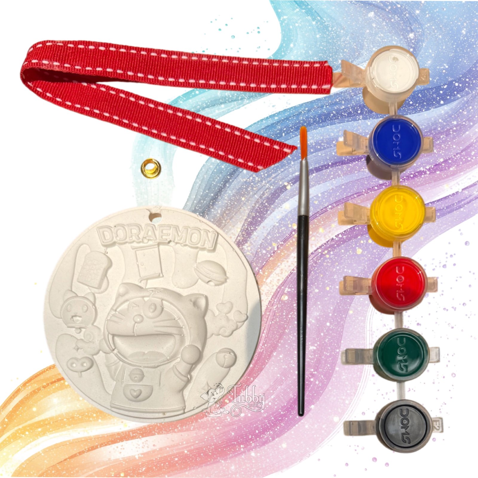 An all-in-one DIY craft hanging kit arranged against a whimsical colorful background. The kit features a white, 4-inch round embossed keepsake plate with a 3D Doraemon character design, a fine-tip paintbrush, a strip of six vibrant acrylic paint pots, a red ribbon with white contrast stitching, and a single golden metal eyelet.