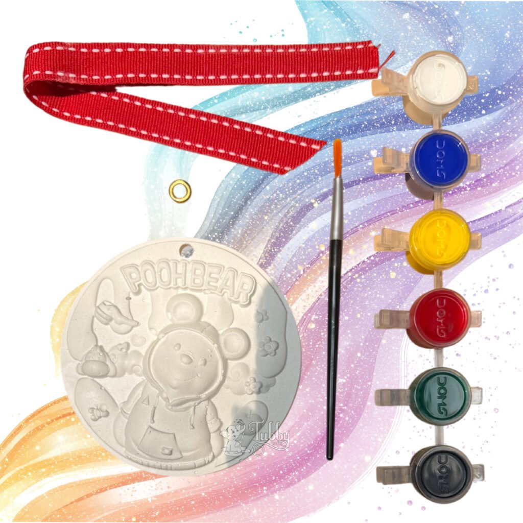 An all-in-one DIY craft kit for children displayed against a colorful, swirly pastel background. The kit features a white, 4-inch round embossed keepsake plate with a 3D design of the cartoon character Winnie the Pooh wearing a hooded sweatshirt under the words "POOH BEAR." The kit also includes a fine-tip paintbrush, a strip of six vibrant acrylic paint pots, a red ribbon with white contrast stitching, and a single golden metal eyelet.